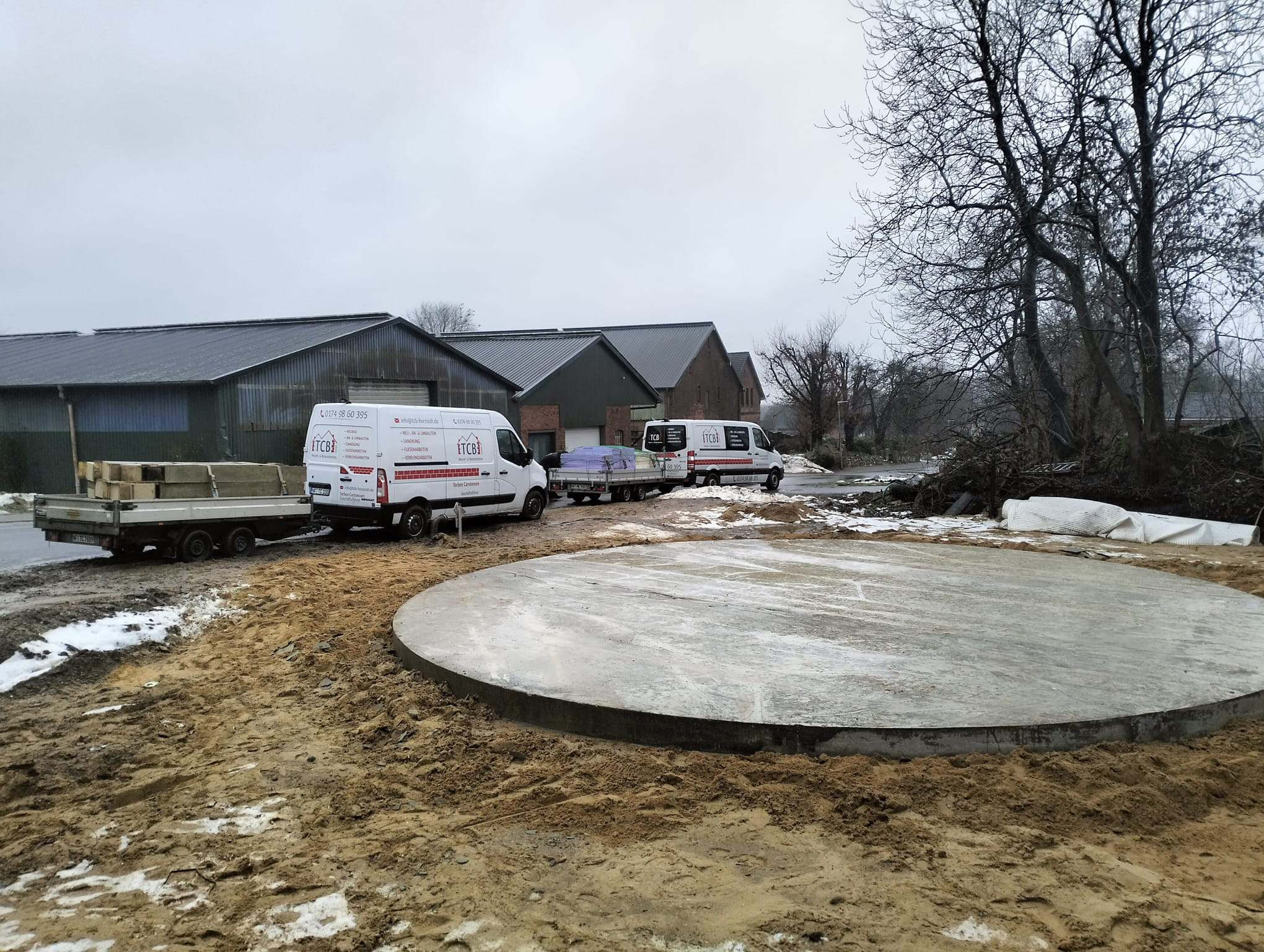 Runde Betonplatte auf einer Baustelle im Winter, mit ITCB-Firmenfahrzeugen und Anhänger mit Baumaterialien im Hintergrund